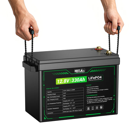 A large black HQST 12.8V 330Ah LiFePO4 battery with green label details, being lifted by two hands holding black straps against a plain white background.